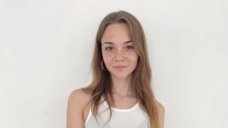 AI-generated close portrait of Mila Hayes, a virtual creator: a smiling young woman with freckles, long light-brown hair and a gold hoop earring, in front of a plain white wall.